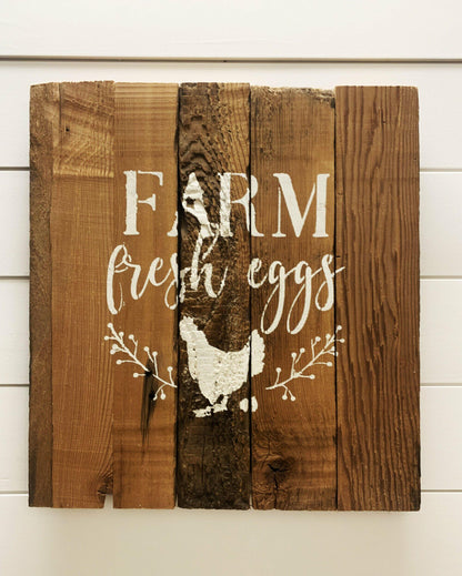 stamped paint design centered on natural and reclaimed barnwood strips put together to create a sign. Design reads Farm Fresh Eggs with a chicken laying an egg underneath underneath and simple florals on either side of the chicken. Wood has distressed characteristics.