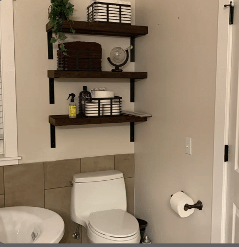 three reclaimed banrwood wall shelves shown in the early american finish. The shelves are shown spaced evenly apart mounted above a toilet in the bathroom. Shelves are decorate with storage for towels, toilet paper, and other bathroom items.