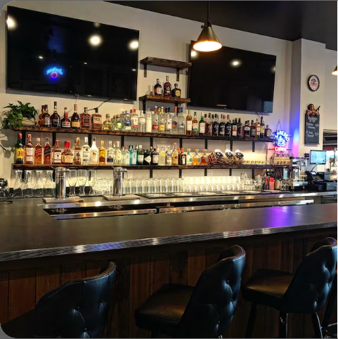 four reclaimed barn wood shelves shown in the oil finish. Shelves are displayed in a bar restaurant. Shelves are lined with bottles of liquor and run the length of the wall. 