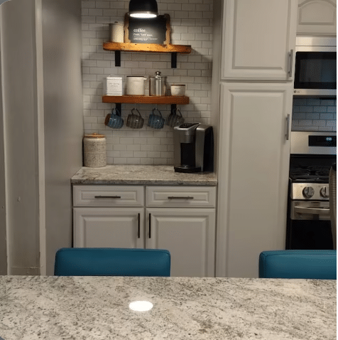 two reclaimed barnwood wall shelves shown in a kitchen on a subway tile backsplash in a coffee knook. Shelves are in the oil finish highlighted by a light above and holding coffee mugs and supplies.