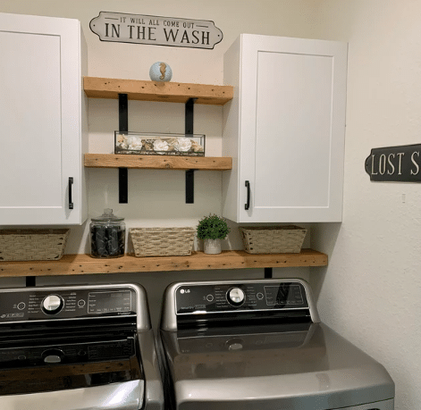 three reclaimed barnwood wall shelves shown in a laundry room. One shelf runs slightly above the washer and dryer with the others in between two cabinets. The shelves are shown in the natural finish with nail holes proudly displayed along the face of the shelves.