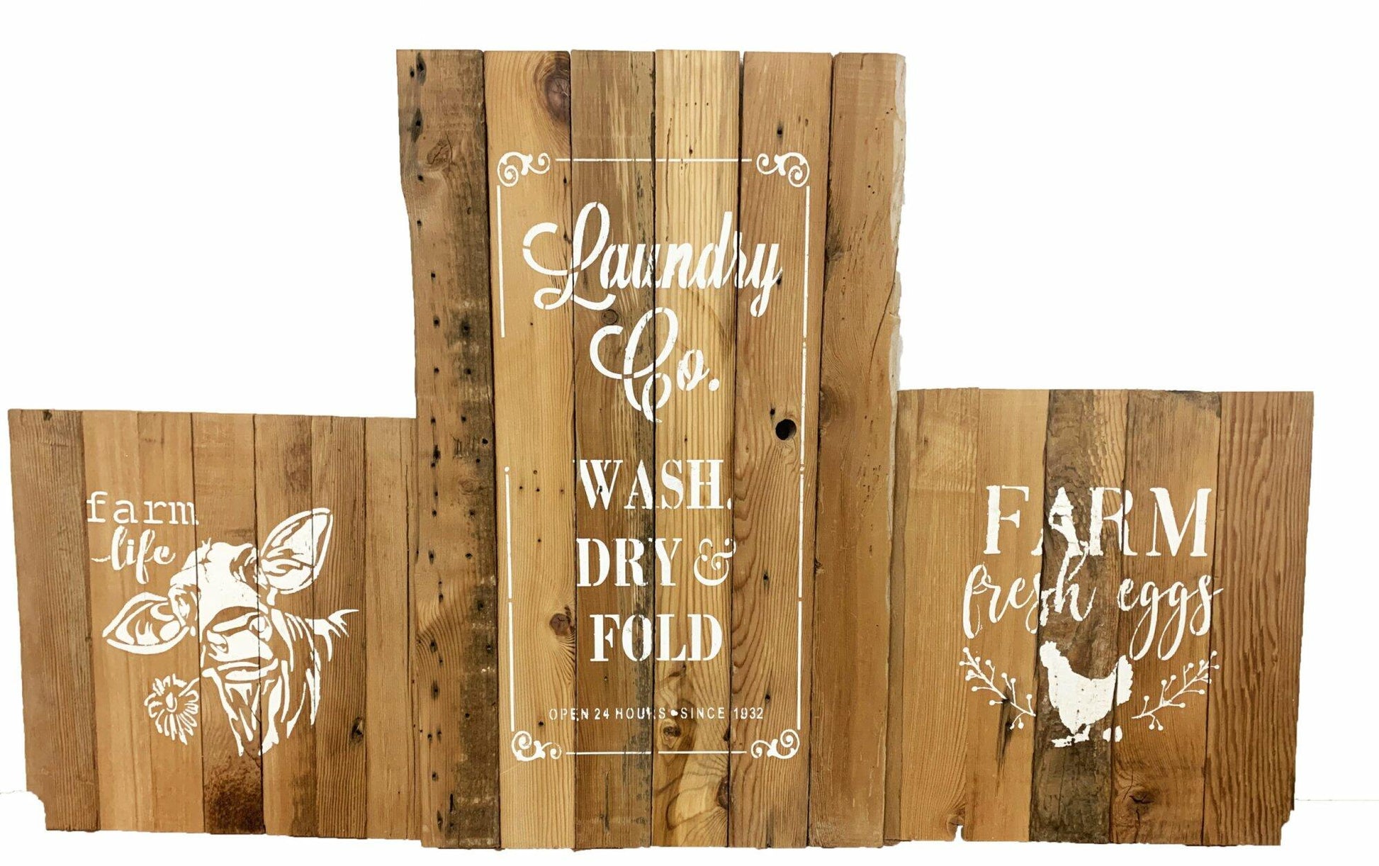 three reclaimed barnwood signs, two smaller to the left and right, one larger in the middle. Each sign has a stamped paint design on them. On sign to the left it reads Farm Life with a cow holding a flower in its mouth underneath. The large middle sign reads Laundry Co. Wash, Dry, & Fold. Open 24 hours since 1932. Smaller sign to the right reads Farm Fresh Eggs with a chicken laying an egg underneath and florals on each side of chicken. Wood has distressed characteristics.