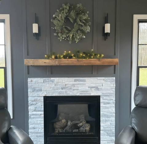 a reclaimed wood floating fireplace mantel in the natural option. The mantel is mounted on a dark wall above a fireplace, providing both contrast and cohesion. The knots and wood grain are prevalent in this mantel.