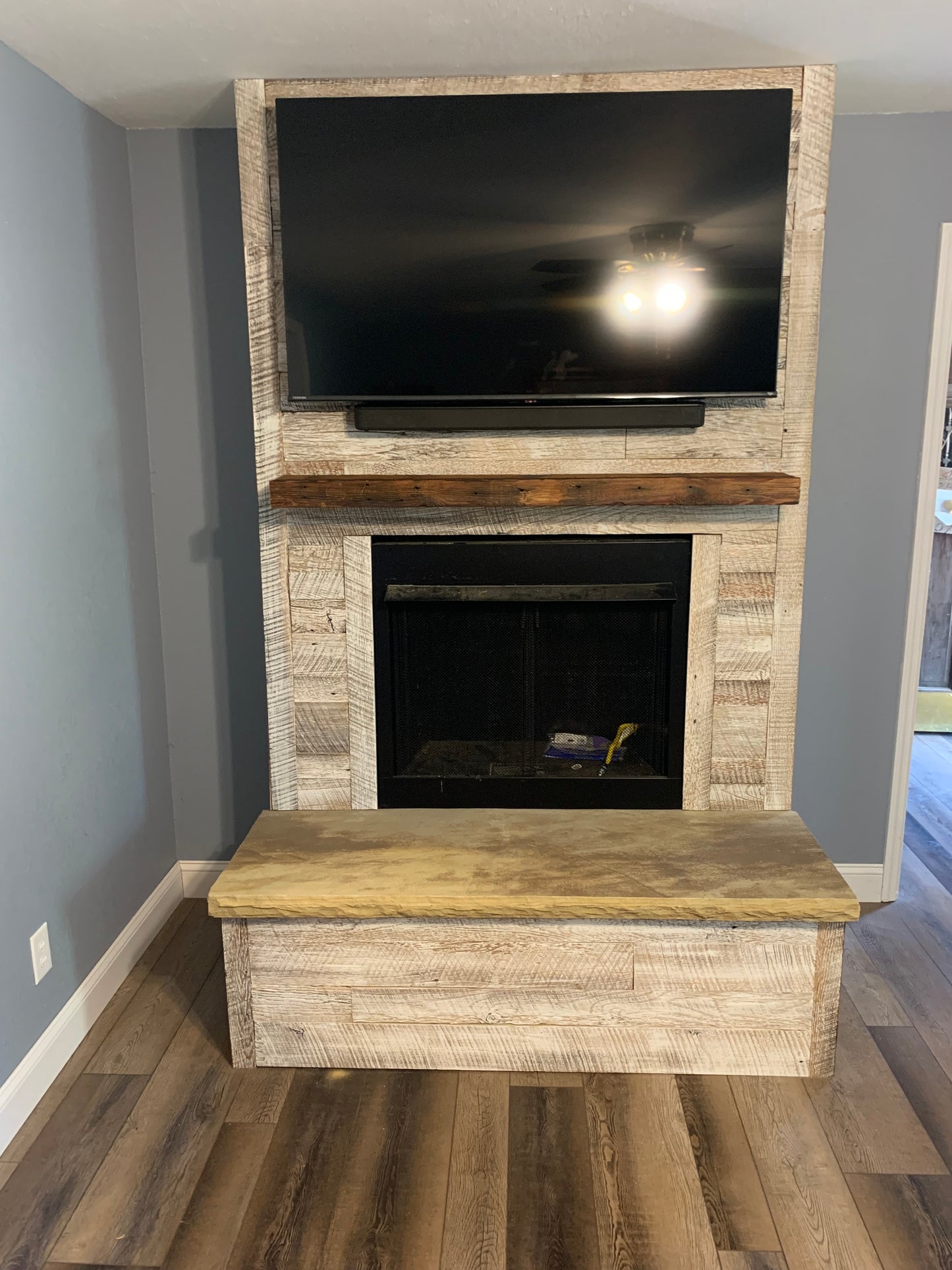 antique white reclaimed barnwood planks used to wrap a fireplace jut out. TV mounted towards the top with a reclaimed, oiled mantel underneath, and a fireplace opening under the mantel. Wood is brown with a white wash finish applied.