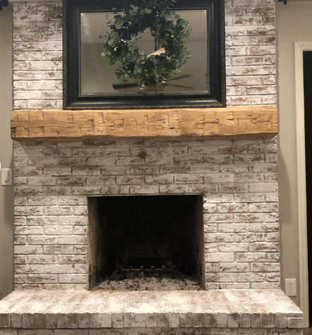 natural wood colored, hand hewn beam showing the woods grain patterns on a white washed, brick fireplace. Mantel has prominent axe marks across the front.