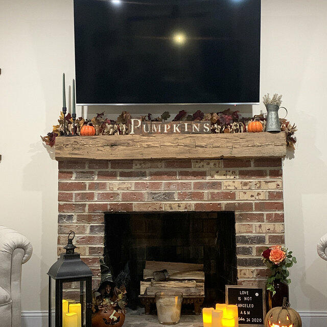 natural wood colored, hand hewn beam showing the woods grain patterns and knots on a traditional brick fireplace. Mantel has light axe marks across the front with knots, and displays a natural characteristic called checking which is similar to cracking. Decorated for fall or Halloween.