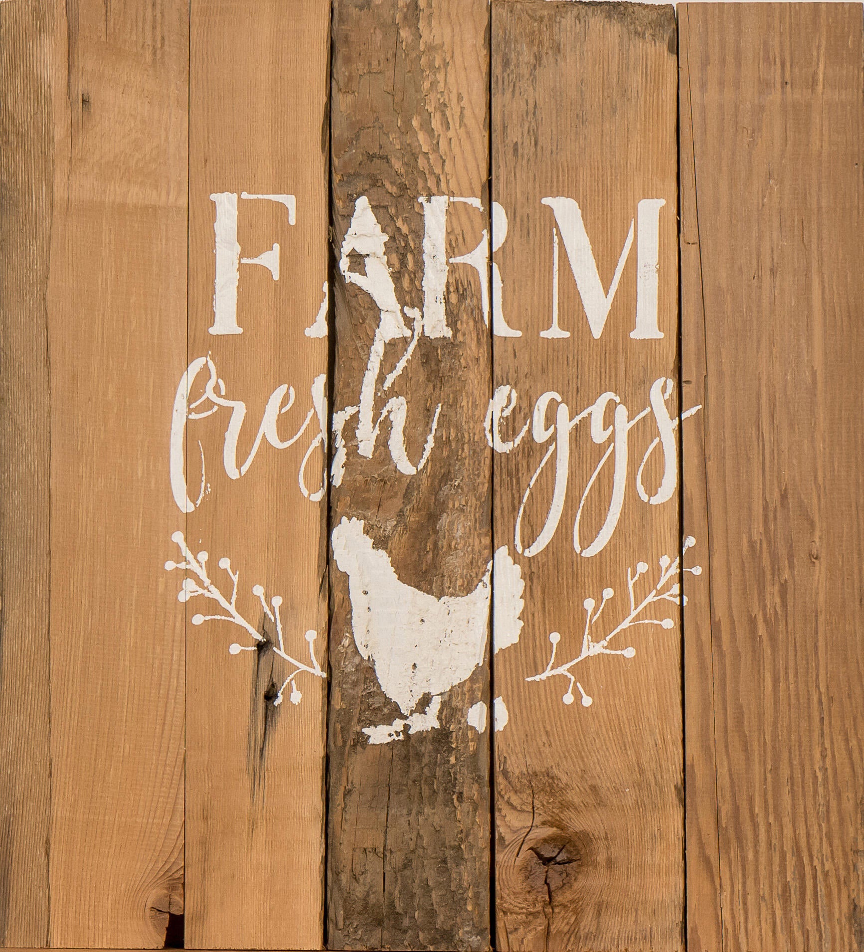 close up of stamped paint design centered on natural and reclaimed barnwood strips put together to create a sign. Design reads Farm Fresh Eggs with a chicken laying an egg underneath underneath and simple florals on either side of the chicken. Wood has distressed characteristics.