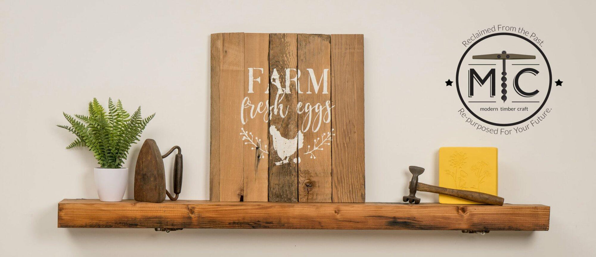 stamped paint design centered on natural and reclaimed barnwood strips put together to create a sign. Design reads Farm Fresh Eggs with a chicken laying an egg underneath underneath and simple florals on either side of the chicken. Wood has distressed characteristics. Shown on a skip planed, oiled mantel with decor items on either side.