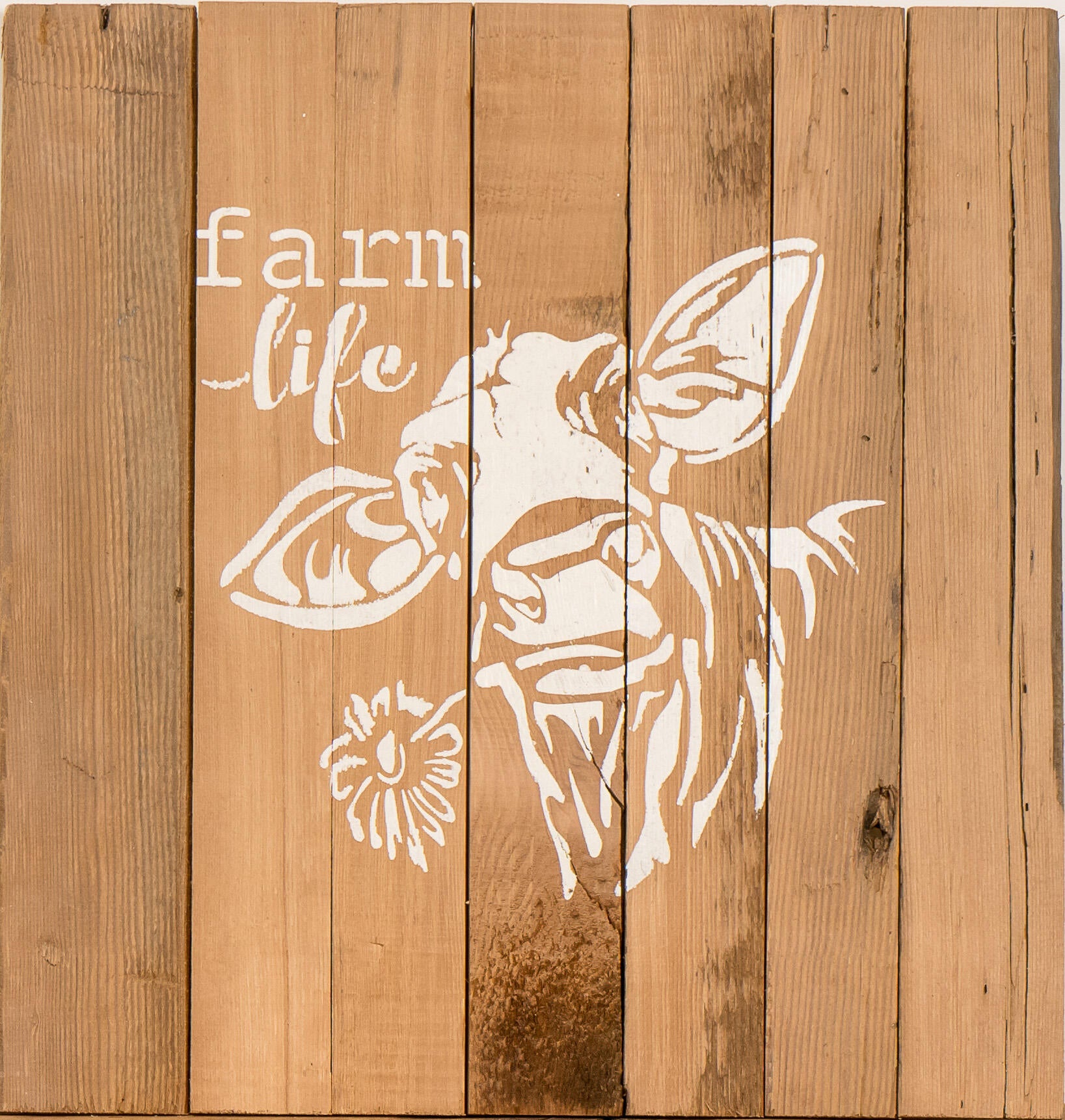 close up of stamped paint design centered on natural and reclaimed barnwood strips put together to create a sign. Design reads Farm Life with a cow holding a flower in its mouth underneath. Wood has distressed characteristics.