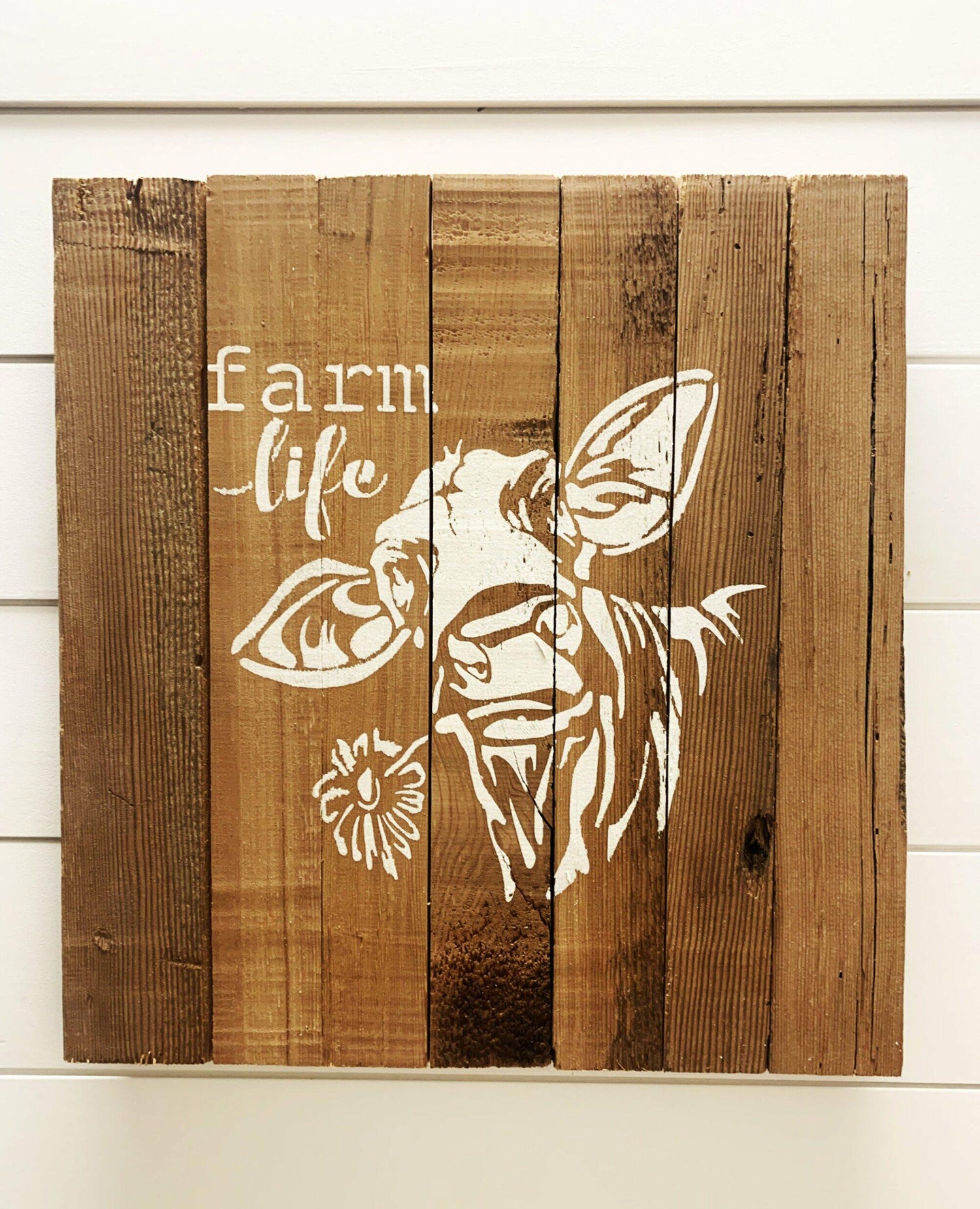stamped paint design centered on natural and reclaimed barnwood strips put together to create a sign. Design reads  Farm Life with a cow holding a flower in its mouth underneath. Wood has distressed characteristics.