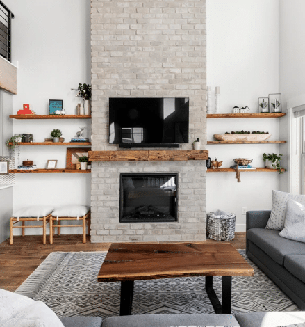 natural wood colored, hand hewn beam showing the woods grain patterns on a light brick fireplace. Mantel shows natural characteristic called checking which is similar to cracking and mortise pockets which are rectangular openings. Hand hewn mantel is flanked by four, 2" thick shelves.