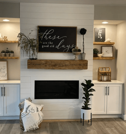 natural wood colored, hand hewn beam showing the woods grain patterns on a light shiplap fireplace. Mantel shows natural characteristic called checking which is similar to cracking. Hand hewn mantel is flanked by four, 2" thick shelves over cabinets.