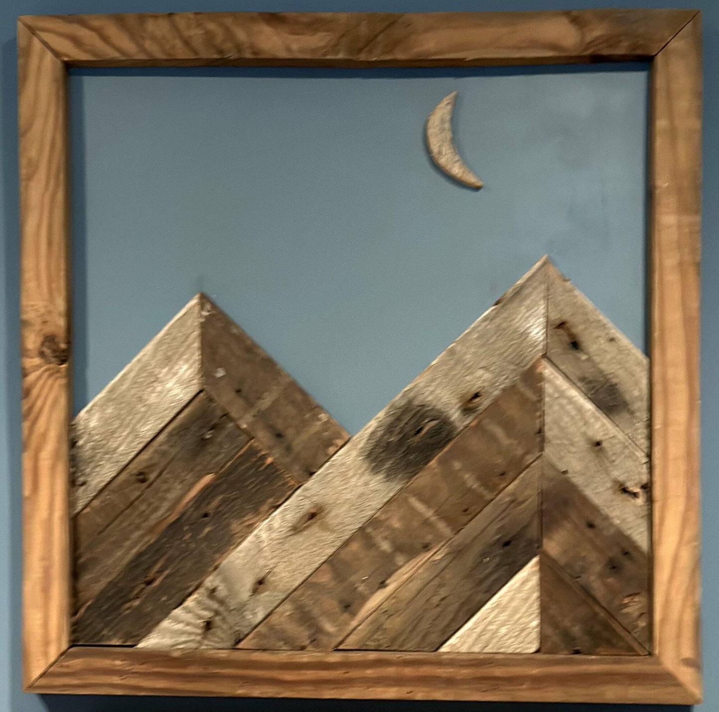 wall decor that looks like two mountains with a crescent moon overtop. Mountains, moon, and frame of wall decor all made from reclaimed barnwood. Wood shows grain patterns and other distressed characteristics.
