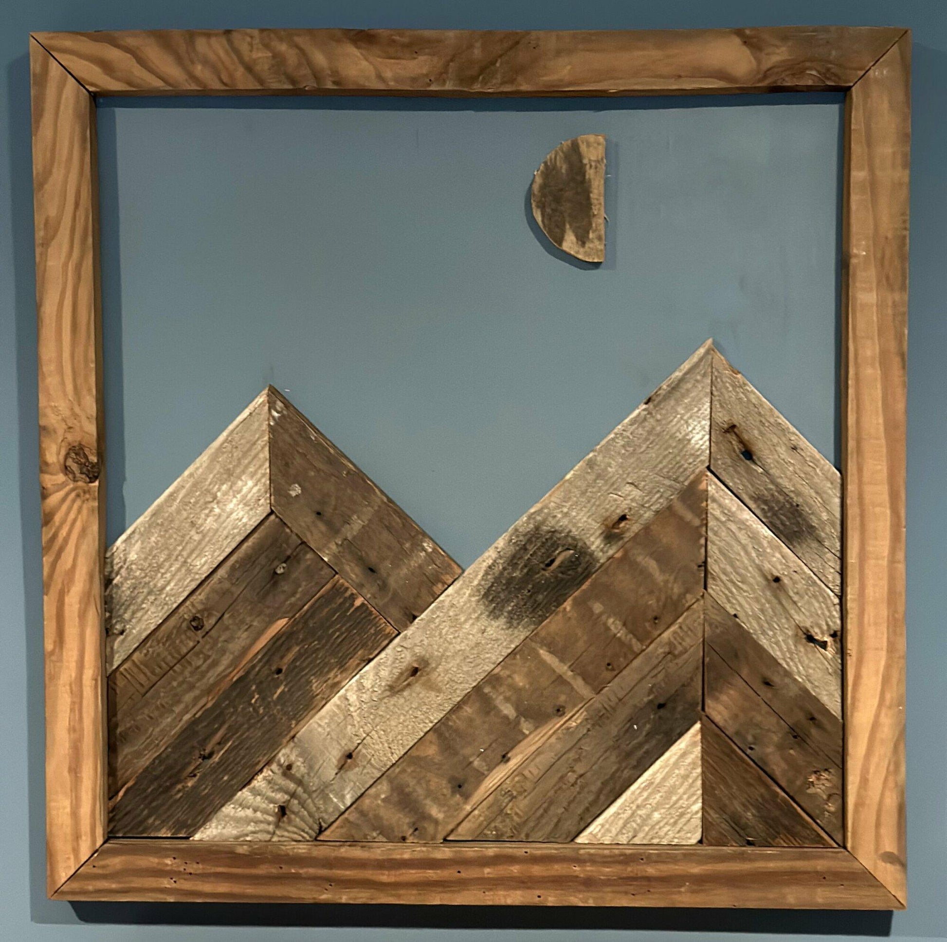 wall decor that looks like two mountains with a moon half full overtop. Mountains, moon, and frame of wall decor all made from reclaimed barnwood. Wood shows grain patterns and other distressed characteristics.