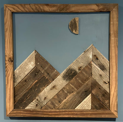 wall decor that looks like two mountains with a moon half full overtop. Mountains, moon, and frame of wall decor all made from reclaimed barnwood. Wood shows grain patterns and other distressed characteristics.
