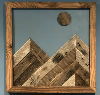 wall decor that looks like two mountains with a full moon overtop. Mountains, moon, and frame of wall decor all made from reclaimed barnwood. Wood shows grain patterns and other distressed characteristics.