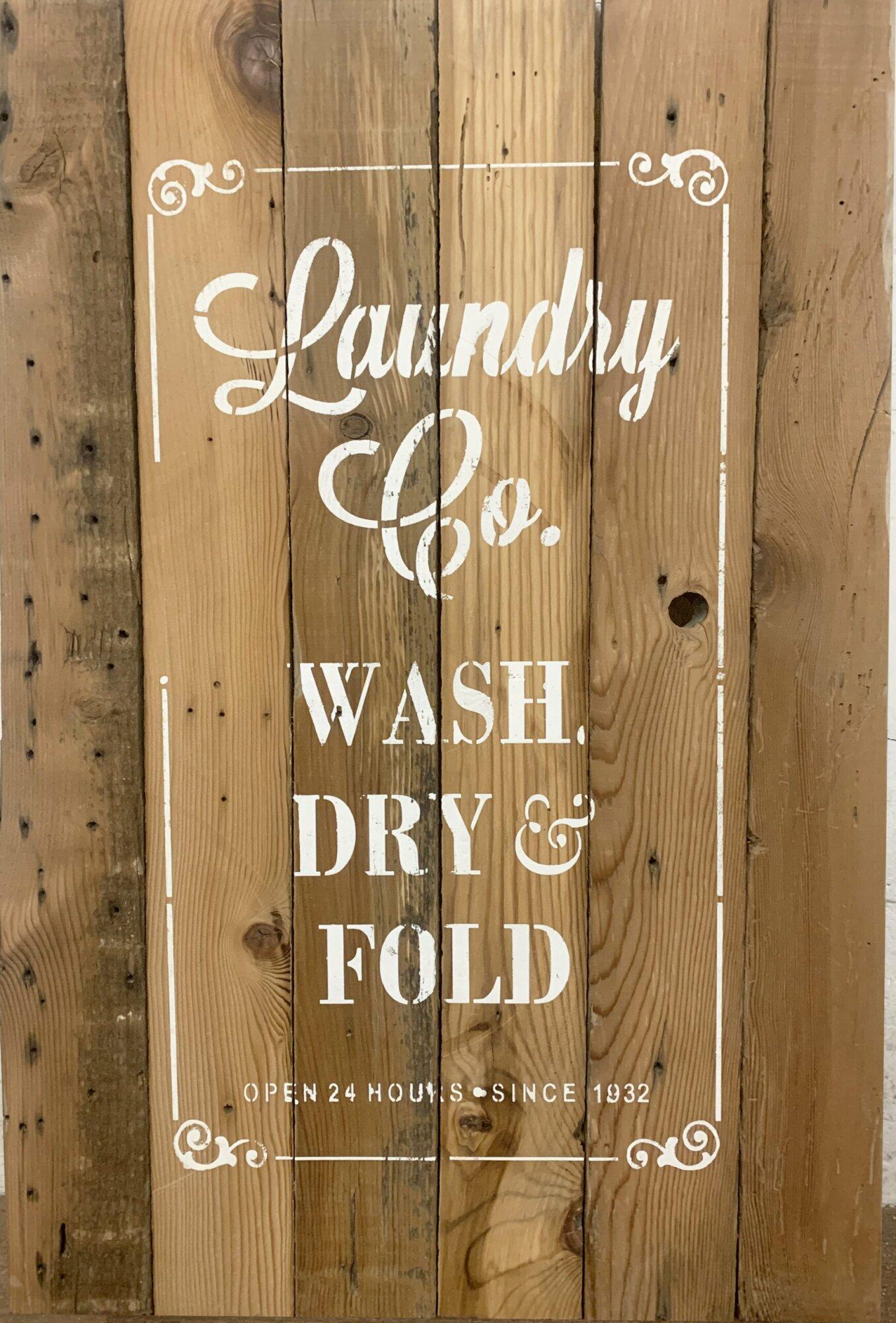 close up of stamped paint design centered on natural and reclaimed barnwood strips put together to create a sign. Design reads Laundry Co. Wash, Dry, & Fold. Open 24 hours since 1932. Text on sign is outlined with scroll design on corners and simple lines. Wood has distressed characteristics.