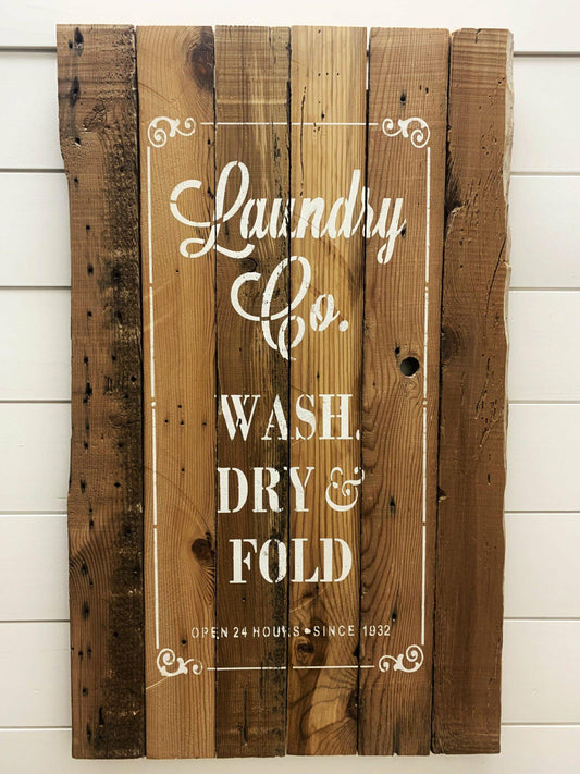 stamped paint design centered on natural and reclaimed barnwood strips put together to create a sign. Design reads  Laundry Co. Wash, Dry, & Fold. Open 24 hours since 1932. Text on sign is outlined with scroll design on corners and simple lines. Wood has distressed characteristics.