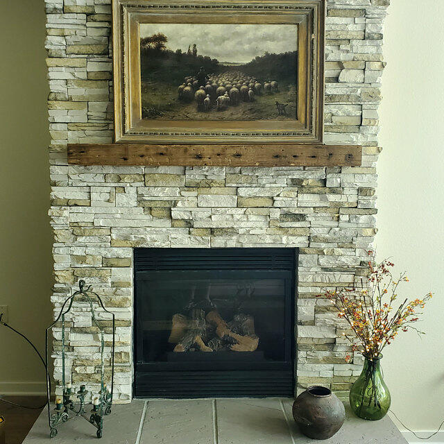 a reclaimed wood floating fireplace mantel in the natural option. Mantel is proudly displayed on a white stone fireplace with nail holes displayed across the face of the mantel.