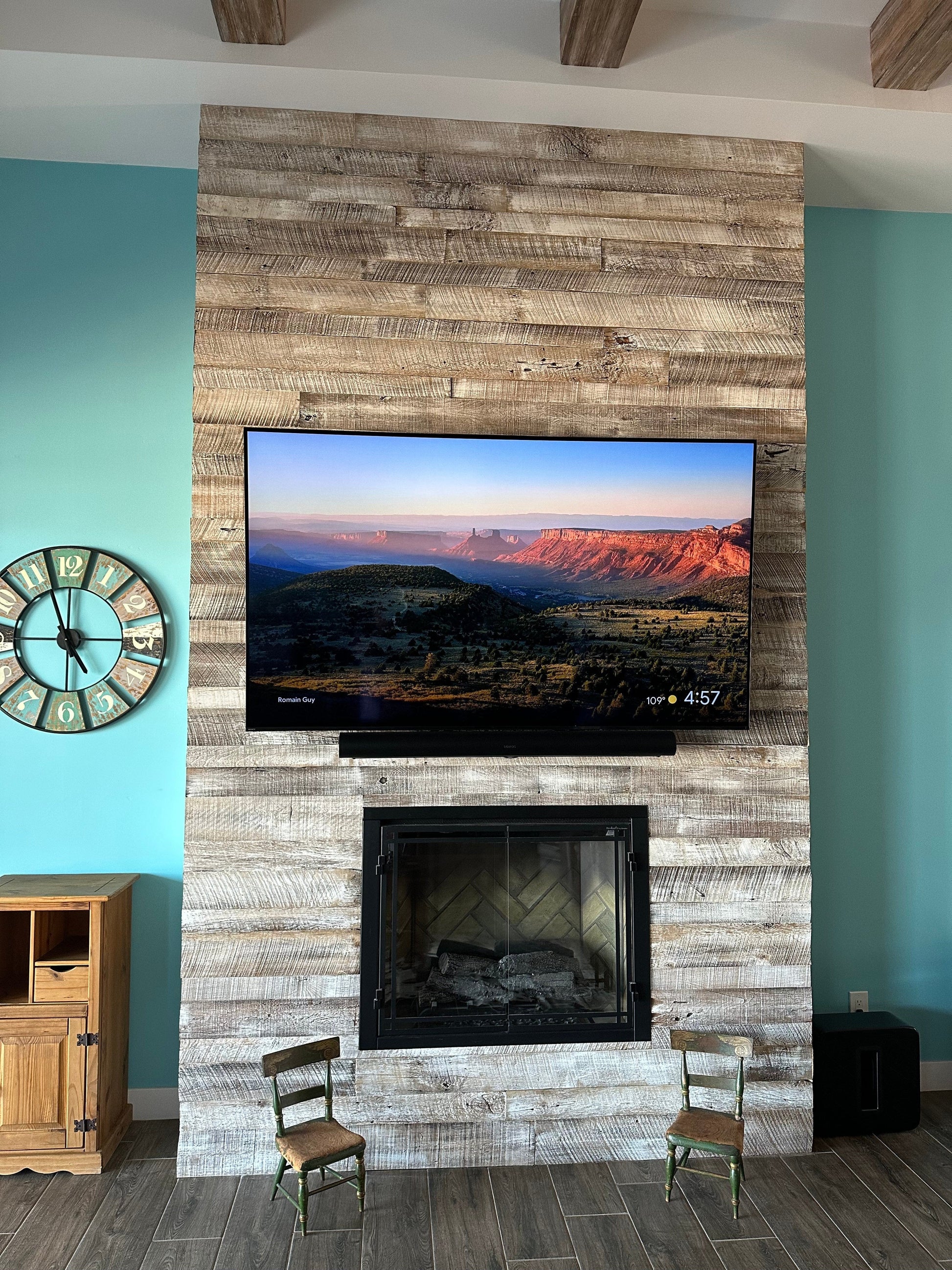 antique white reclaimed barnwood planks used to wrap a fireplace jut out. TV mounted towards the top with fireplace opening under the television and two small chairs for decor. Wood is brown with a white wash finish applied.