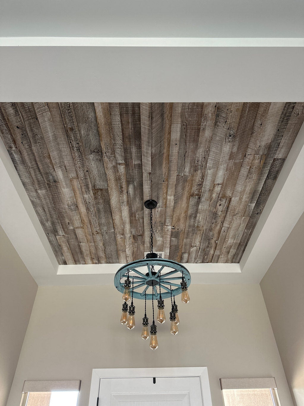 antique white reclaimed barnwood planks used on the ceiling of a foyer. Wood is brown with a white wash finish applied. A rustic wheel chandelier hangs from the middle with Edison bulbs hanging from the wheel.