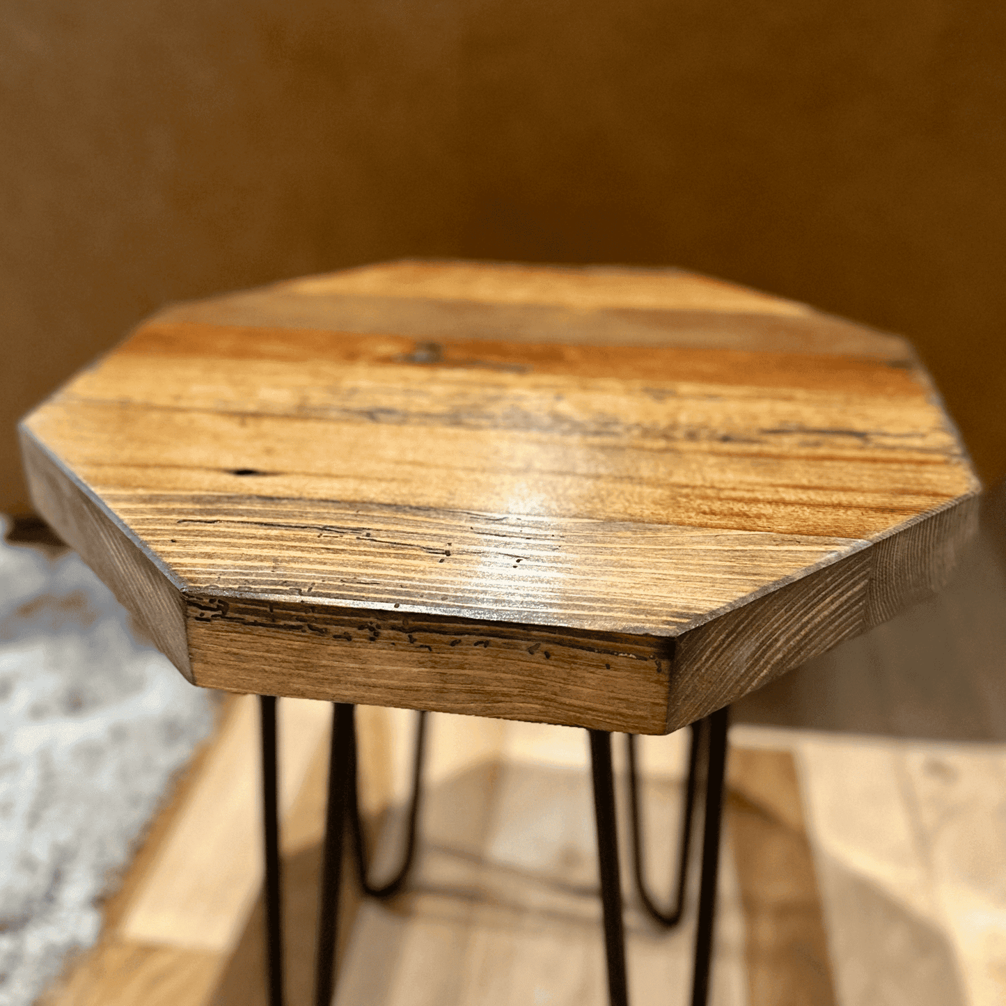 a reclaimed wood table in an octagonal shape and early american finish. The table is supported by four hairpin legs and is shown next to a couch as a side table.