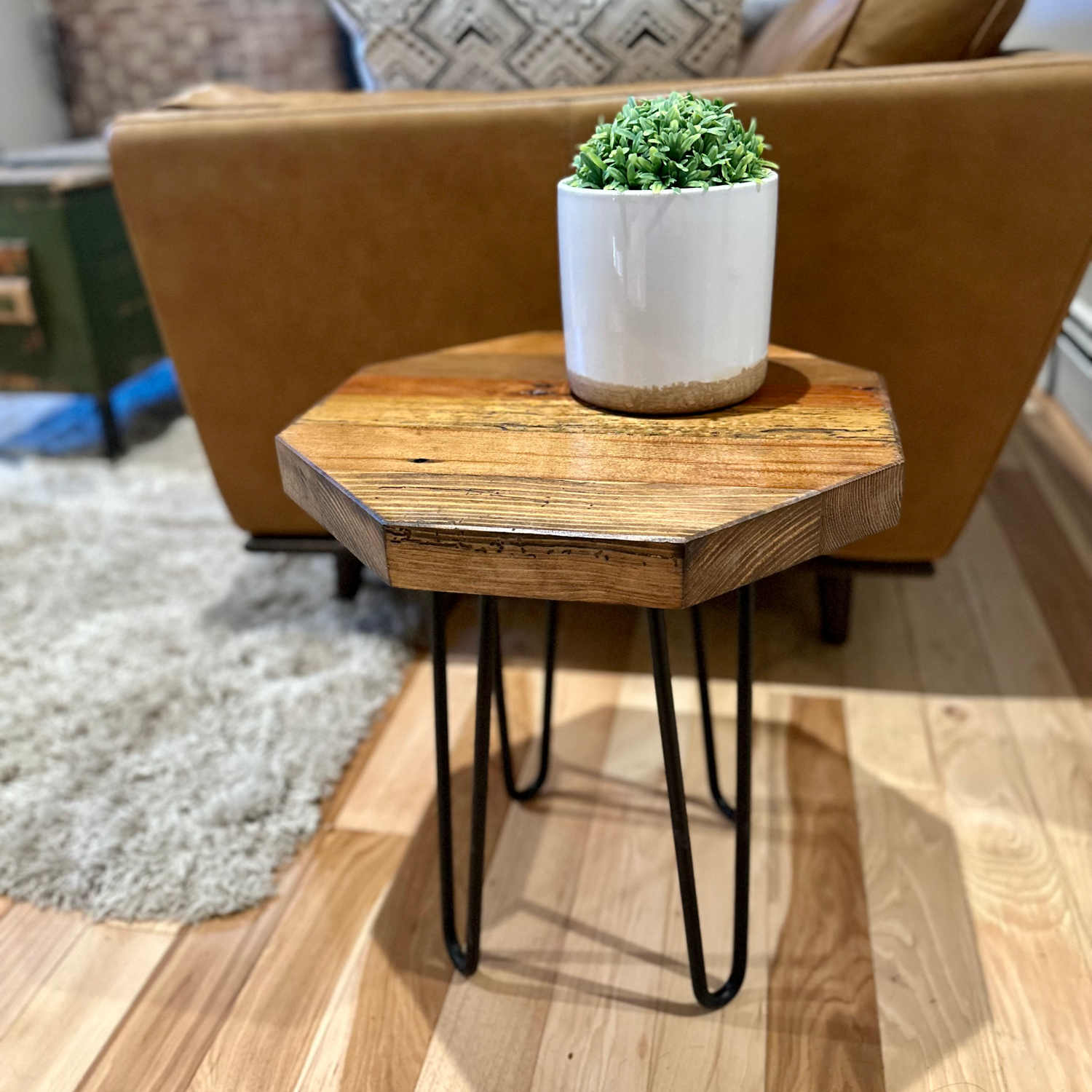 a reclaimed wood table in an octagonal shape and early american finish. The table is supported by four hairpin legs and has a small plant displayed on top. The table is shown next to a couch as a side table.