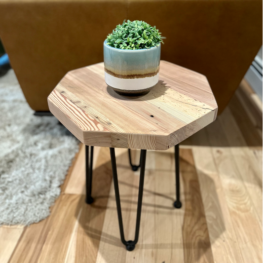 a reclaimed wood table in an octagonal shape and unfinished finish. The table is supported by four hairpin legs and has a small plant displayed on top. The table is shown next to a couch as a side table.