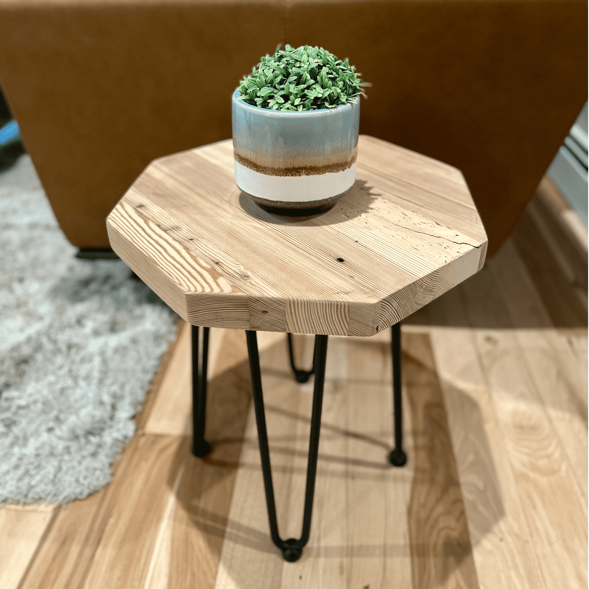 a reclaimed wood table in an octagonal shape and unfinished finish. The table is supported by four hairpin legs and has a small plant displayed on top. The table is shown next to a couch as a side table.