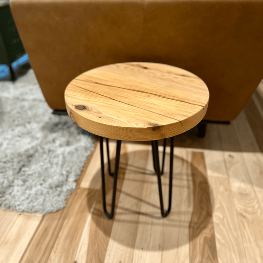 a reclaimed wood table in an round shape and sealed unfinished finish. The table is supported by four hairpin legs and is shown next to a couch as a side table.