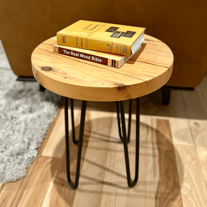 a reclaimed wood table in an round shape and sealed unfinished finish. The table is supported by four hairpin legs and is shown next to a couch as a side table with books on top for decor.
